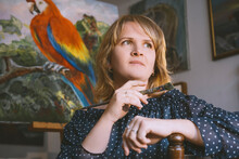 Thoughtful Woman With Hand On Chin Holding Paintbrush In Front Of Bird Painting At Workshop