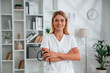 © standret - Holding stethoscope. Young female doctor in white coat is indoors