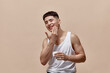 © master1305 - Portrait of smiling, handsome, young guy with clean, spotless face applying face moisturizing cream against light brown studio background. Concept of male beauty, skincare, cosmetology, men's health