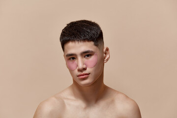 Portrait of handsome young asian guy taking care after skin, applying under eye patches against light brown studio background. Concept of male beauty, skincare, cosmetology, men's health