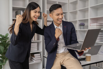 Happy young Asian businessman and woman shaking hands to congratulate successful collaboration on business deal with documents and laptop on table in the office.