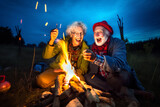 Fototapeta Sport - Old couple of husband and wife, enjoying retirement - they are warming up by a campfire at blue hour while on a trip. Generative AI
