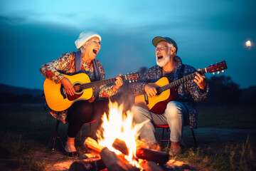 Wall Mural - Old couple of husband and wife, enjoying retirement - they are singing and playing guitar by a campfire at blue hour while on a trip. Generative AI