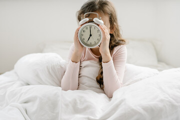 8 years girl holding alarm clock 7 am hidding the face, sitting on bed under white blanket, sleep at morning. Time go to school, wake up early at home