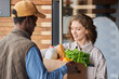 © Seventyfour - Waist up portrait of young woman receiving grocery delivery outdoors and holding bag with fresh vegetables
