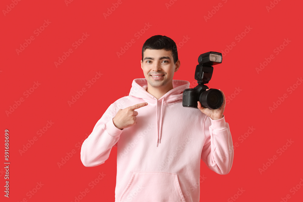 Young male photographer pointing at professional camera on red background