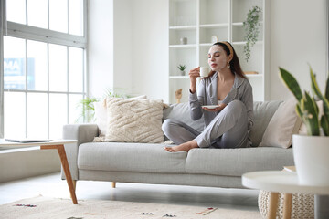 Wall Mural - Young woman with sheet mask and cup of coffee sitting on sofa at home