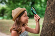 © _KUBE_ - Children's education. Portrait of a smiling little girl scout in a straw hat looks at the tree bark through a magnifier. The concept of curious childhood
