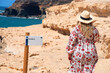 © Safi - Turista rubia de pelo largo con un vestido de flores y un sombrero de paja junto a un cartel que dice 'Sendero' en los acantilados de la costa de Fuerteventura en un día soleado en las Islas Canarias