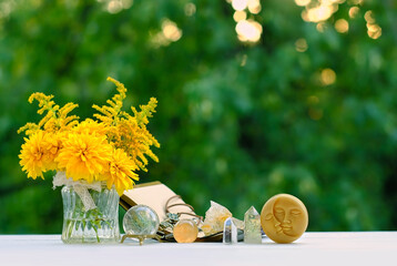 Wall Mural - Flowers, witchcraft items, book and crystals on table close up, blurred green natural background. Wiccan, Slavic magic practice. Witchcraft, esoteric spiritual ritual. Mystic, divination, occultism