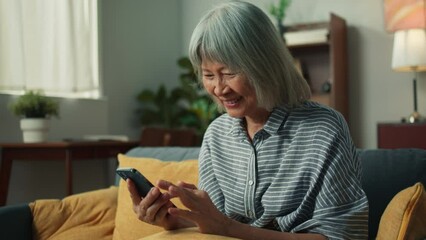 Wall Mural - Happy Asian elderly woman smile holding mobile phone checking email received good news, feeling surprised excited. Cheerful aged female looking at smartphone screen sit on sofa in home living room.