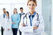 © ronstik - portrait of female doctor wearing white coat standing in hospital hallway with staff in background. generative AI