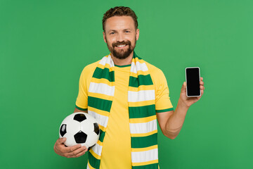 Wall Mural - cheerful man in striped scarf holding football and smartphone with blank screen isolated on green.