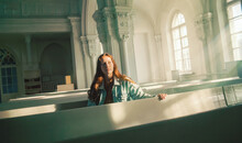 A Beautiful Young Red-haired Woman Is Sitting On Old Pews Inside A Protestant Church, Illuminated By The Radiance Of The Sun's Rays And Divine Light. Faith And Religion.