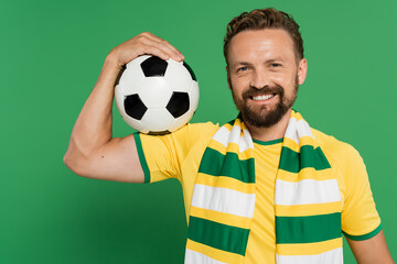 Wall Mural - smiling soccer fan in striped scarf and yellow t-shirt holding football isolated on green.