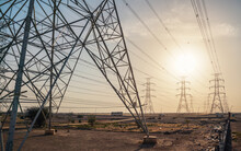 Large Tall Steel Power Pylons In Rather Flat Landscape Of Saudi Arabia, Only Black Silhouettes Visible Against Bright Afternoon Sun
