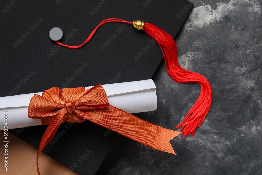 Diploma with red ribbon, graduation hat and book on dark grey table