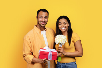 Cheerful millennial black guy gives bouquet and gift box to woman, congratulating with birthday, anniversary