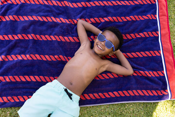 African american shirtless boy with hands behind head wearing sunglasses and lying on towel