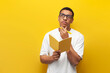 © Богдан Маліцький - pensive guy african american student in white t-shirt and glasses writes in notebook and plans on yellow background