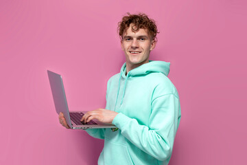 curly guy in mint hoodie is holding laptop and typing on pink isolated background, man in sweatshirt is using computer