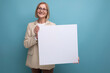© Ivan Traimak - smiling mature woman in jacket demonstrating blank paper mockup on studio background with copy space
