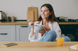 © ViDi Studio - Young fun smiling european happy housewife woman wear casual clothes look aside eat breakfast muesli cereals with milk fruit in bowl cooking food in light kitchen at home alone. Healthy diet concept.