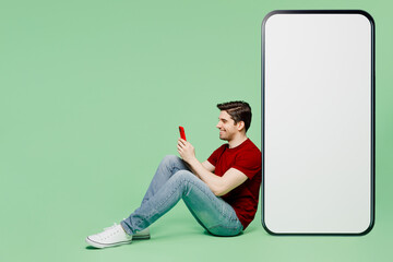 Wall Mural - Full body side view young man he wears red t-shirt casual clothes sit near big huge blank screen mobile cell phone with workspace area using smartphone isolated on plain pastel light green background.
