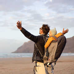 Wall Mural - Father rising hands to the sky while enjoying pure nature carrying his infant baby boy son in backpack on windy sandy beach of Famara, Lanzarote island, Spain at sunset. Family travel concept