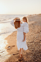 Wall Mural - A small child, a girl in her mother's arms, both in white dresses, walk barefoot along the ocean at sunset.