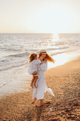 Wall Mural - A small child, a girl in her mother's arms, both in white dresses, walk barefoot along the ocean at sunset.