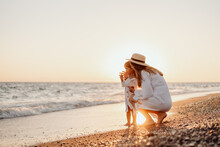 A Young Mother Sat Down And Holds Her Little Daughter By The Hand On The Ocean At Sunset. Girls In White Dresses And Long Hair.