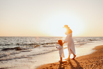 Wall Mural - A young mother holds her little daughter by the hand and together they walk along the ocean towards the sunset. Girls in white dresses and with long hair that develops the wind
