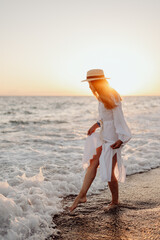 Wall Mural - A young, beautiful girl with long hair in a straw hat and a white dress stands in the water on the ocean at sunset. Blurred background. wave crashes on the shore
