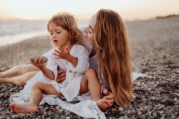 Wall Mural - A young mother and daughter in white dresses are sitting on a rocky ocean shore at sunset. Little daughter looks at the stone in her hands
