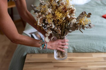 Canvas Print - Hands placing a vase of dry flowers on the wooden table in a cozy bedroom