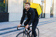 © makedonski2015 - A smiling male courier rides a bicycle along a modern building in the city. A delivery man with a yellow backpack
