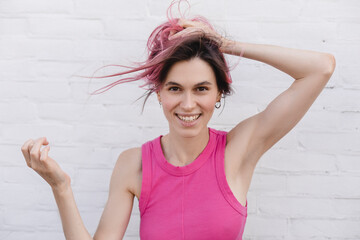 Sticker - Portrait of young pink hair woman in pink top, plays with hair, arms raised up, photo on light brick background. Girl smiling, look playful, posing, make hairstyle.