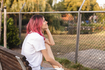 Poster - Woman with pink hair sitting on bench and smiling, walking in park, looking carefree and happy. Modern girl breathing fresh air on a walk, look free and joyful.