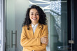 © Liubomir - Portrait of successful business woman inside office, latin american boss in yellow suit smiling and looking at camera, mature female worker with curly hair standing with arms crossed.
