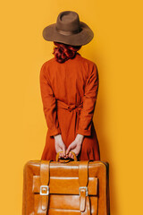 Back side view on stylish redheaded woman in hat and brown dress with suitcase on yellow background