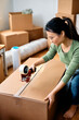 © Drazen - Asian woman sealing cardboard boxes with tape dispenser while relocating in apartment.