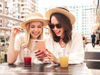 © halayalex - Two young beautiful smiling hipster female in trendy summer white dress and hats. Sexy carefree women drinking fresh vegetable cocktail smoothie drink in plastic cup with straw. Using phone apps,