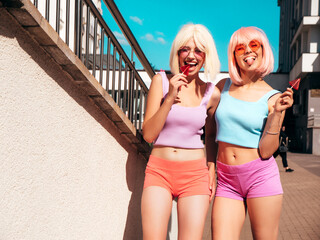 Wall Mural - Two young beautiful smiling hipster female in trendy summer clothes. Sexy carefree women posing in the street in wigs. Positive funny models having fun at sunset. In sunglasses. Eating candy lollipop