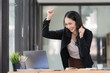 © Songsak C - Successful young Asian business woman achieving goals excited raised hands rejoicing with laptop in the office.
