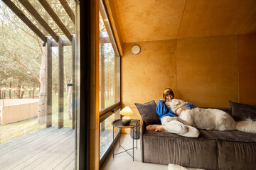 Canvas Print - Woman cares her dog while sitting on a couch by the window, spending leisure time together in wooden house on nature