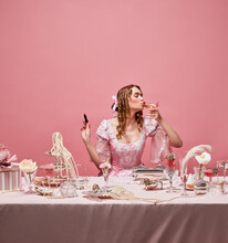 Shot Of Adorable Blond Princess Wearing Fancy Pink Dress And Sitting At Beautifully Laid Table With Wine Glass And Lipstick Over Studio Background. Drinking Cocktail