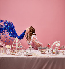 Medieval attractive blond queen wearing fancy pink dress sitting at laid table with hangover over studio background. Alcohol party