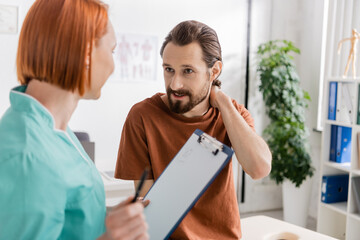 Wall Mural - bearded man touching painful neck near blurred physiotherapist with clipboard in consulting room.