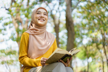 Wall Mural - Happy cheerful Asian beautiful Muslim woman reading a book - Koran under the tree. Female muslim student studying an Islam Koran.